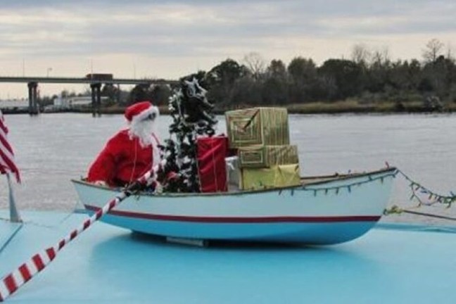 Santa figure in a small boat with gifts and a tree on a river with bridge in background.