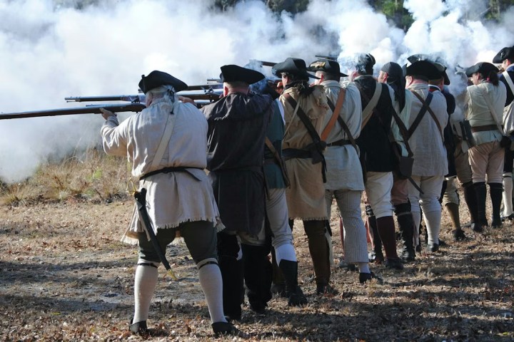 Historical reenactors firing muskets in a line, with smoke in the background.