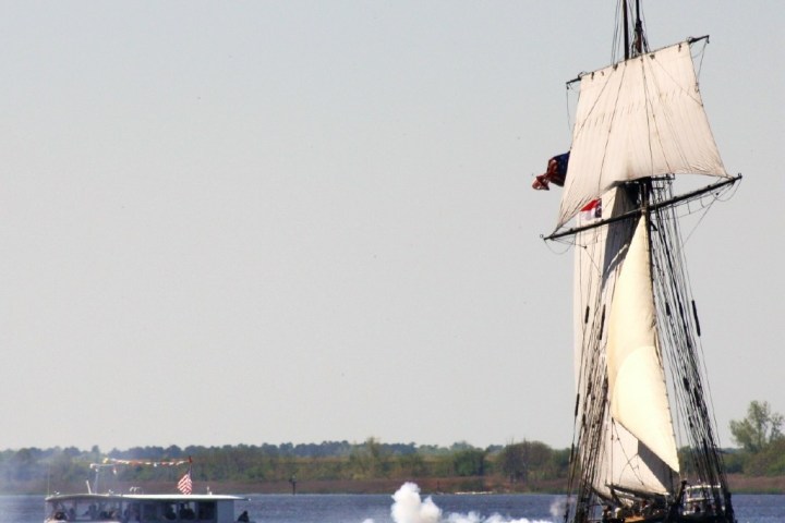 Tall ship firing cannon near a modern boat on a calm body of water.