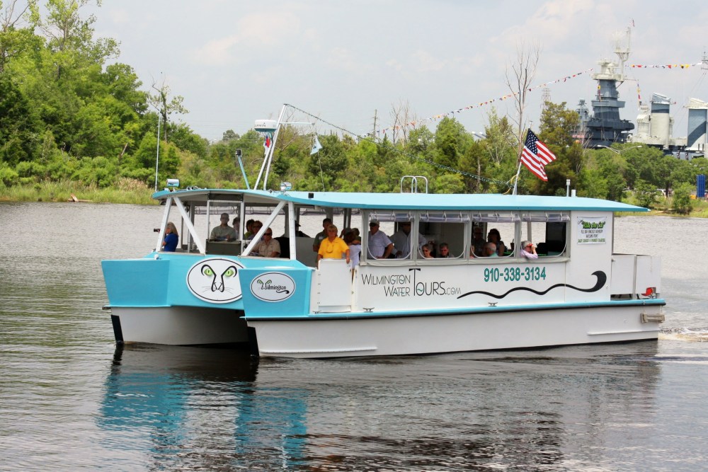 Tour boat with passengers on a river, US flag flying, trees in background.